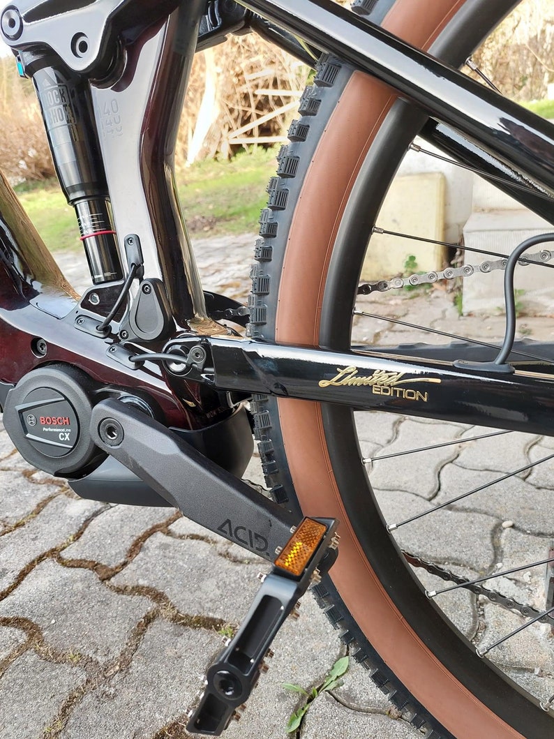 Puede incluir: Bicicleta de monta&ntilde;a el&eacute;ctrica negra con neum&aacute;tico marr&oacute;n, motor Bosch negro y plateado y pedal negro con reflector amarillo. La bicicleta tiene un cuadro negro con una inscripci&oacute;n dorada "Limited Edition".