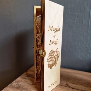 May include: A wooden box with a hinged lid, engraved with the names "Megija & Elvijs" and the date "19.07.2023". The box is made of light wood with a dark brown stain around the edges. The box is open and the inside is lined with brown paper.