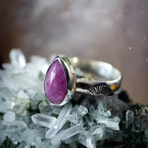 May include: A silver ring featuring a teardrop-shaped pink gemstone set in a bezel setting. The ring is resting on a bed of clear quartz crystals.