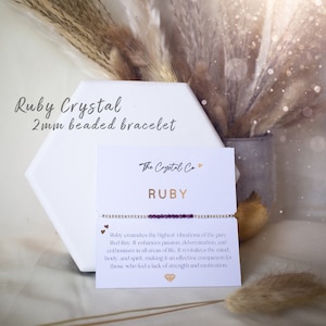 May include: A purple crystal bracelet with gold beads and a card that reads "Ruby Crystal 2mm beaded bracelet The Crystal Co RUBY Ruby emanates the highest vibrations of the pure Red Ray. It enhances passion, determination, and enthusiasm in all areas of life. It revitalizes the mind, body, and spirit, making it an effective companion for those who feel a lack of strength and motivation."