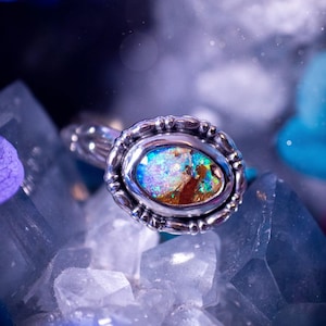 May include: An oval-shaped ring with a silver-colored setting and a colorful opal stone. The opal displays a mix of blues, greens, and oranges. The ring is displayed on a bed of clear crystals, with purple and blue floral accents.