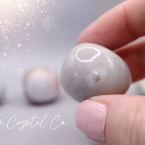 May include: A close-up of a smooth, white agate stone with a grey and white pattern. The stone is being held by a hand with pink nail polish. The stone is a tumbled stone, which means it has been smoothed by rubbing against other stones.
