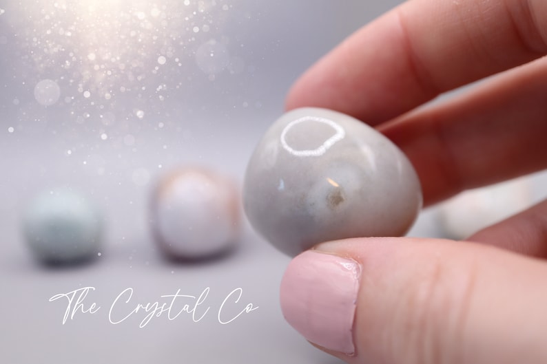 May include: A close-up of a smooth, white agate stone with a grey and white pattern. The stone is being held by a hand with pink nail polish. The stone is a tumbled stone, which means it has been smoothed by rubbing against other stones.