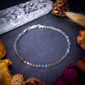 Könnte beinhalten: Ein zartes Armband aus silberner Kette mit schillernden Labradoritperlen. Das Armband liegt auf einer dunkelgrauen Oberfläche mit einem klaren Quarzkristall und getrockneten Blumen.