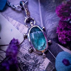 May include: A silver pendant necklace with a large, faceted, emerald-green gemstone. The pendant is set in a silver bezel with decorative details. The necklace is displayed on a background of feathers, purple flowers, and vintage paper.