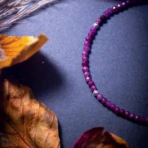 Può includere: Un braccialetto di perline di pietre preziose viola con accenti argentati su uno sfondo grigio scuro con petali di fiori secchi.