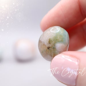 May include: A round, green, white and brown agate gemstone with a smooth, polished surface. The gemstone is being held in a hand.
