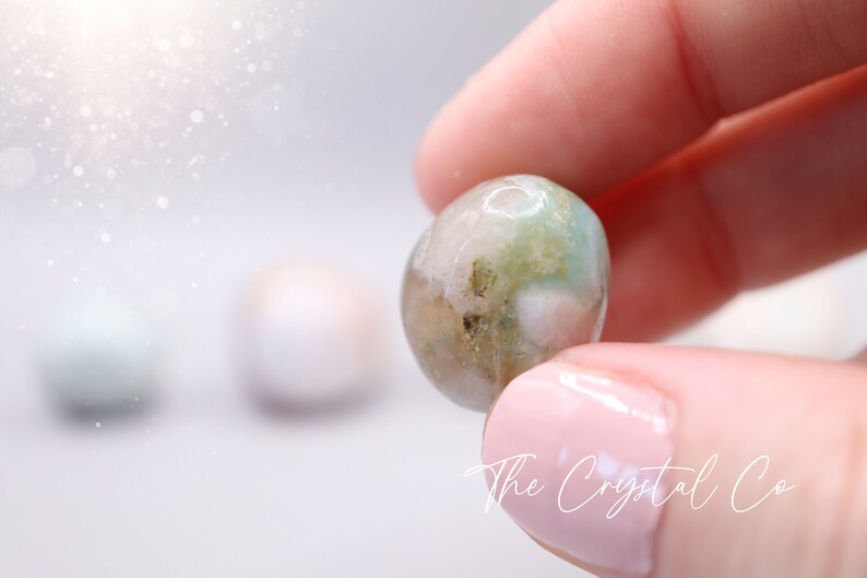 May include: A round, green, white and brown agate gemstone with a smooth, polished surface. The gemstone is being held in a hand.