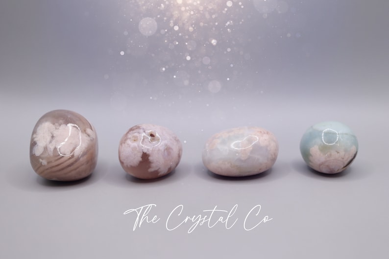May include: Four smooth, polished agate stones in shades of pink, white, and blue. The stones are arranged in a row on a grey surface. The Crystal Co. is written in white script below the stones.