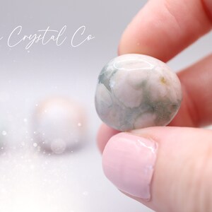 May include: A close-up of a green and white agate sphere, with a swirling pattern of white and green. The sphere is being held in a hand with pink nail polish.