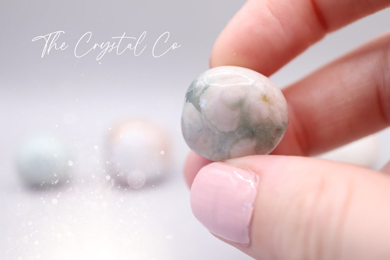 May include: A close-up of a green and white agate sphere, with a swirling pattern of white and green. The sphere is being held in a hand with pink nail polish.