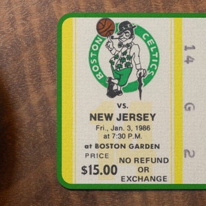 May include: A vintage Boston Celtics ticket stub for a game against the New Jersey team on January 3, 1986 at Boston Garden. The ticket is green and yellow with the Boston Celtics logo and the text "SEC. ROW SEAT 14 G 2". The price of the ticket is $15.00.
