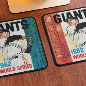 May include: Two square coasters with a vintage design featuring a baseball player in a Giants uniform. The coasters have a red, white, and blue color scheme and the text "Giants 1962 World Series".