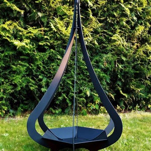 Może przedstawiać: Czarny metalowy palenisko o unikalnym kształcie łzy. Palenisko ma okrągłą misę i wysoką, zakrzywioną ramę. Znajduje się na trawniku z zielonym żywopłotem w tle.