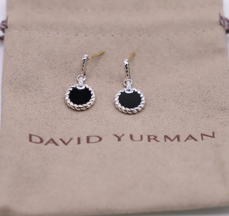 May include: David Yurman earrings featuring black onyx and diamonds.  These drop earrings have a cable-rope design surrounding each circular black onyx stone.  Small diamonds accent the top of each earring. The earrings are displayed on a beige jewelry pouch.