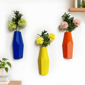 May include: Three geometric wall-mounted vases in solid blue, yellow, and orange colors. Each vase features a faceted, angular design and holds a bouquet of artificial flowers with green leaves and white, yellow, or pink blossoms.