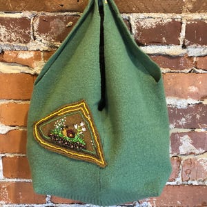 May include: A green knitted tote bag with a brown triangle patch featuring an embroidered snail and flowers.