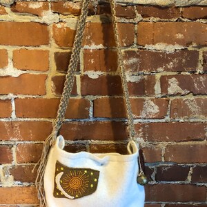 May include: White knitted tote bag with a brown patch featuring an embroidered sun and moon design. The bag has a long, brown braided strap and a small, round, gold-colored charm.