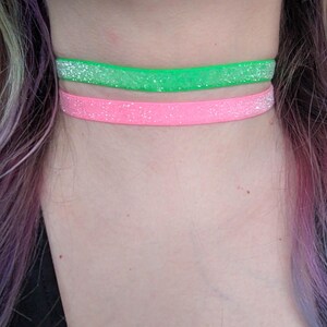 Puede incluir: Dos gargantillas con purpurina, una verde neón y otra rosa, se llevan alrededor del cuello de una persona. Las gargantillas están hechas de un material fino y plano y están cubiertas de purpurina. La persona tiene el pelo multicolor.