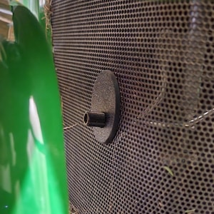 May include: A close-up of a black metal grate with a round, black plastic cap with a small, black metal pipe protruding from it.