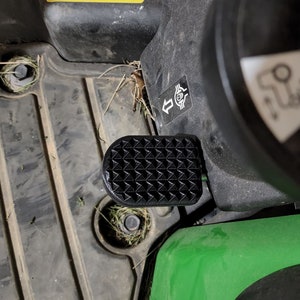 May include: A black foot pedal with a textured surface. The pedal is attached to a green and black machine.