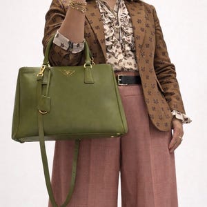 May include: A green leather handbag with a top handle and a shoulder strap. The bag has gold-colored hardware and a Prada logo. The person is wearing a brown blazer with a floral pattern, a patterned blouse, and pink pants.