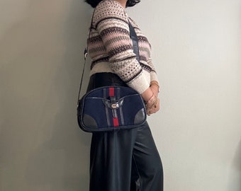 May include: A blue and brown suede shoulder bag with a gold buckle and red and white stripes. The bag is worn by a person wearing a cream and brown striped sweater and black pants.