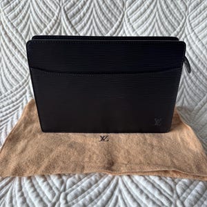 May include: A black rectangular clutch bag featuring a textured horizontal line pattern, a top zipper closure, and a small embossed LV logo in the bottom right corner. The bag is positioned on a tan fabric dust cover with a small LV logo printed on it.
