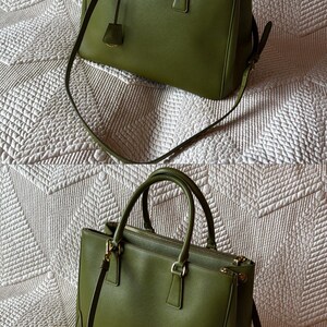 May include: Two olive green handbags with top handles and detachable shoulder straps. The bags have gold-tone hardware and a small triangular logo on the front. The bags are sitting on a white quilted surface.