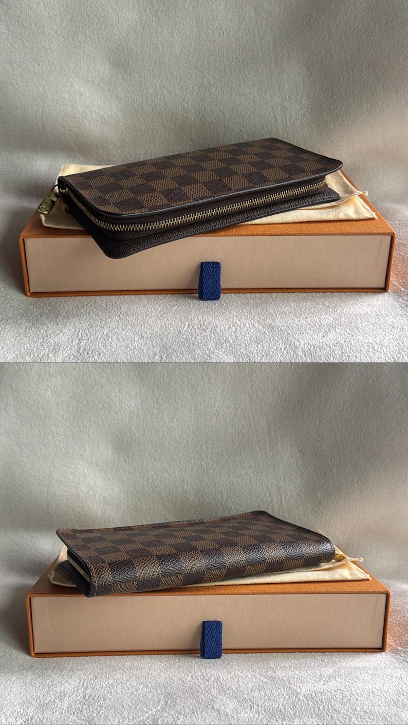 May include: A brown leather wallet with a checkered pattern and a gold zipper. The wallet is in a brown box with a blue ribbon.