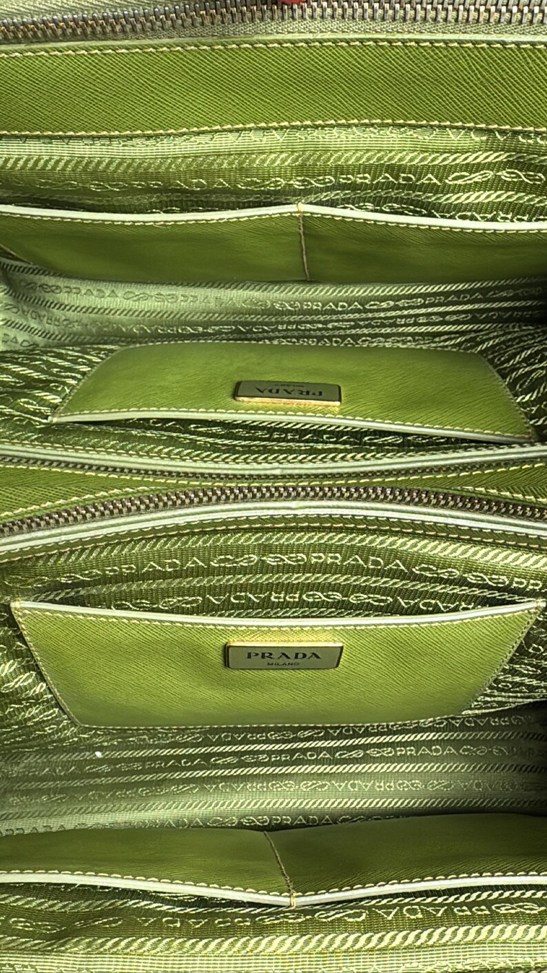 May include: Close-up of a green Prada handbag interior. The bag features multiple compartments with a light green lining and the Prada logo. The interior is detailed with the Prada logo pattern.