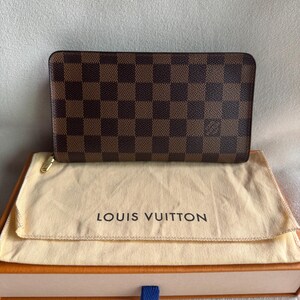 May include: A brown and tan Louis Vuitton wallet with a checkered pattern. The wallet is in a beige dust bag and a brown box with a blue ribbon.