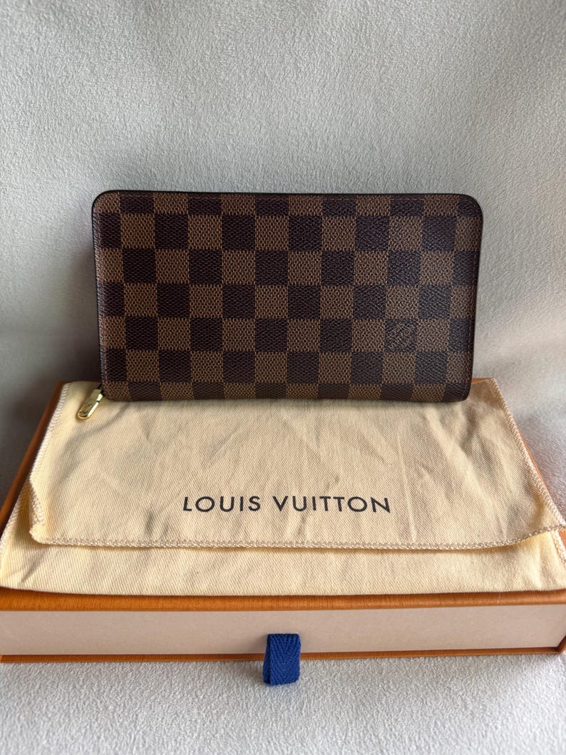 May include: A brown and tan Louis Vuitton wallet with a checkered pattern. The wallet is in a beige dust bag and a brown box with a blue ribbon.