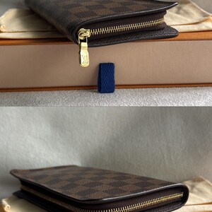 May include: A brown and black checkered leather wallet with a gold zipper and a gold Louis Vuitton logo. The wallet is sitting on a beige box with a blue ribbon.