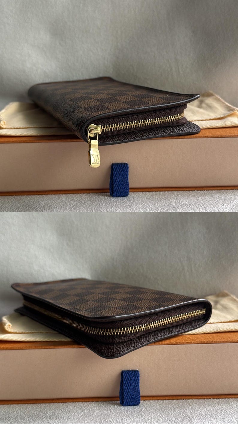 May include: A brown and black checkered leather wallet with a gold zipper and a gold Louis Vuitton logo. The wallet is sitting on a beige box with a blue ribbon.
