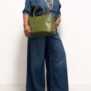 May include: A green leather handbag with gold-colored hardware. The bag has a top handle and a detachable shoulder strap. The person is wearing a denim shirt and wide-leg jeans. The outfit is accessorized with gold jewelry and a pearl necklace.
