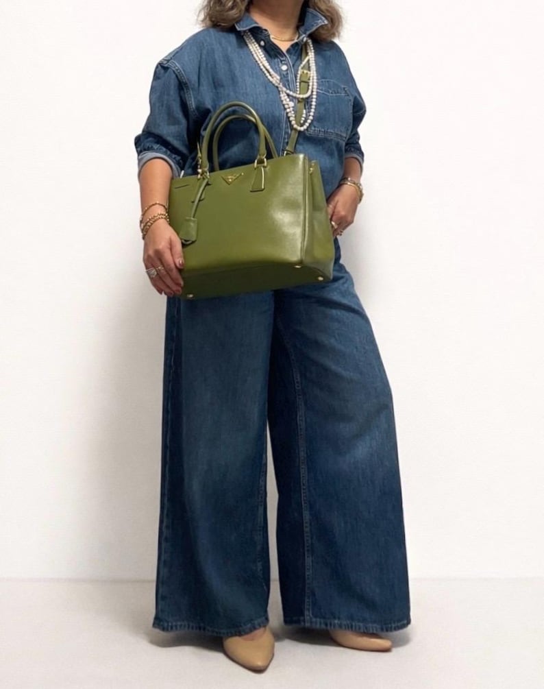 May include: A green leather handbag with gold-colored hardware. The bag has a top handle and a detachable shoulder strap. The person is wearing a denim shirt and wide-leg jeans. The outfit is accessorized with gold jewelry and a pearl necklace.