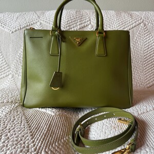 May include: Olive green leather handbag with top handles and a detachable shoulder strap. The bag features gold-tone hardware, including a logo plaque and zipper. The bag is sitting on a white quilted surface.