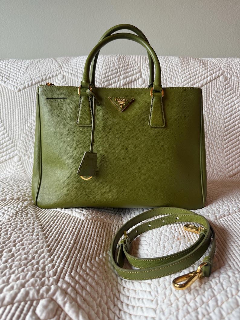 May include: Olive green leather handbag with top handles and a detachable shoulder strap. The bag features gold-tone hardware, including a logo plaque and zipper. The bag is sitting on a white quilted surface.
