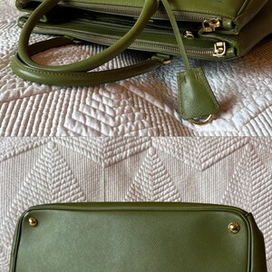 May include: Olive green leather handbag with gold-tone hardware. The bag features double handles, a top zipper closure, and a small leather tag. The bottom of the bag has four gold feet. The bag is sitting on a white patterned surface.