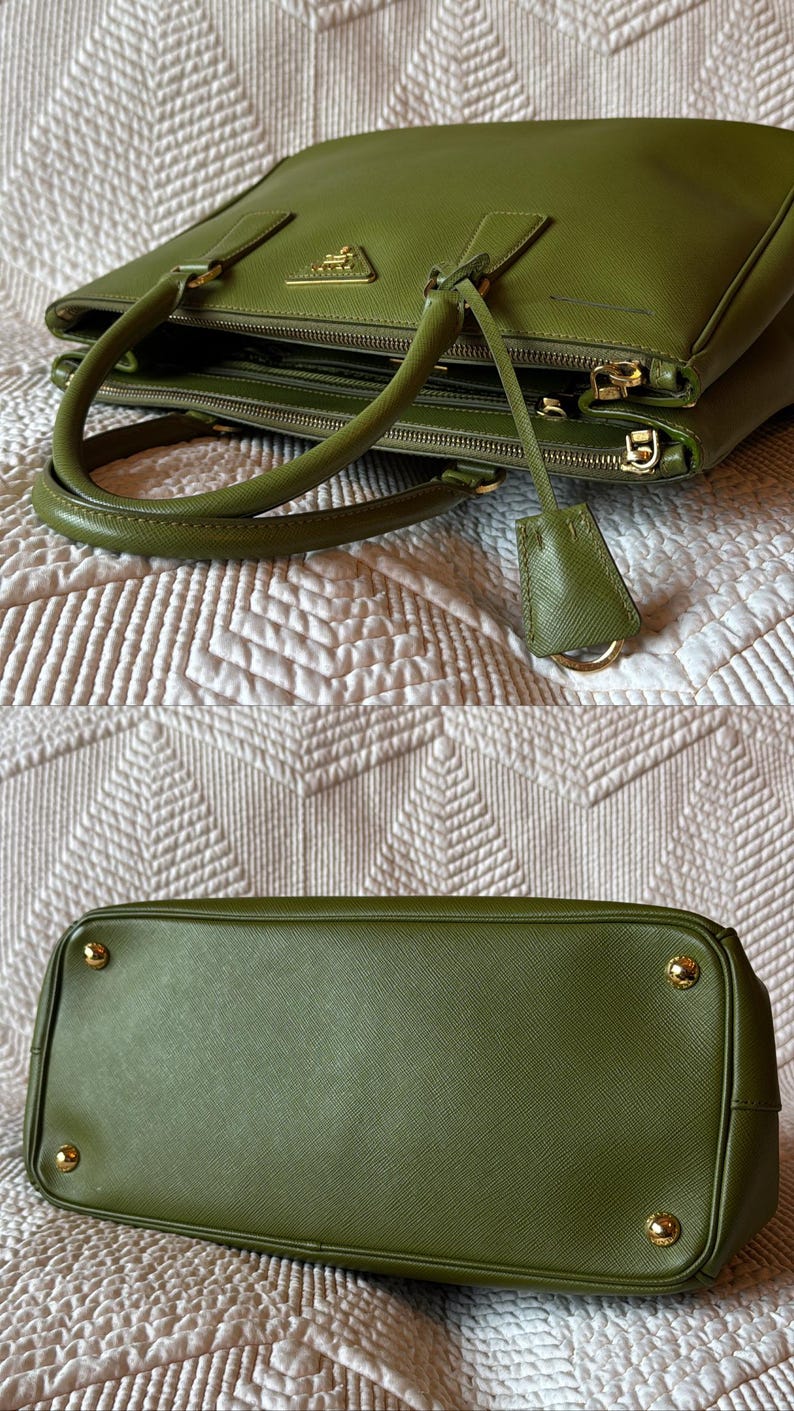 May include: Olive green leather handbag with gold-tone hardware. The bag features double handles, a top zipper closure, and a small leather tag. The bottom of the bag has four gold feet. The bag is sitting on a white patterned surface.