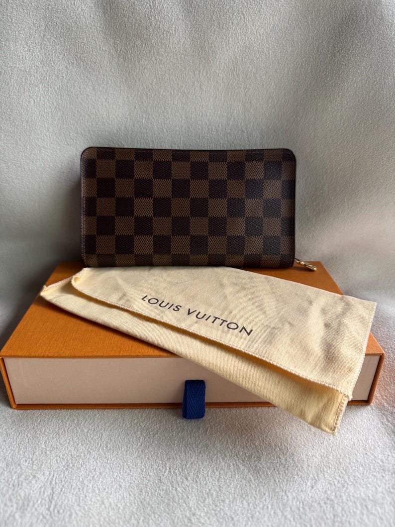 May include: A brown and black checkered Louis Vuitton wallet with a gold zipper pull. The wallet is in a beige dust bag and a brown box with the Louis Vuitton logo.