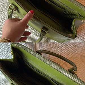 May include: A green leather handbag with gold-toned hardware. The bag has a top handle and a zipper closure. The interior is lined with a matching green fabric and features multiple compartments. The bag is open, revealing the interior.