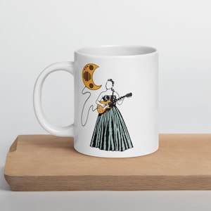 Pode incluir: Caneca de cerâmica branca com uma ilustração em linha de uma pessoa tocando guitarra e um desenho de lua crescente. A pessoa está vestindo um vestido sem alças com listras verticais. A caneca está em uma superfície de madeira.