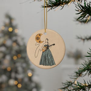May include: Round ceramic ornament with a beige background, featuring a line drawing of a person playing a guitar. The person is wearing a long, striped dress. A crescent moon and musical notes are also depicted. The ornament hangs from a gold string.