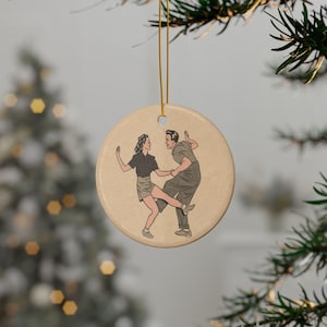 May include: A round ceramic ornament with a vintage illustration of a couple swing dancing. The dancers are depicted in black, gray, and tan tones. The ornament hangs from a gold string, perfect for holiday decor.
