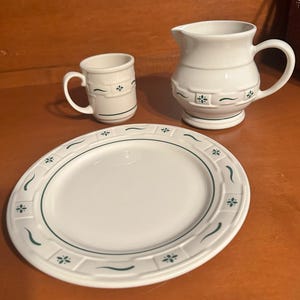 May include: A white ceramic dish set featuring a plate, a mug, and a pitcher. The plate has a green border with floral and curved line designs. The mug and pitcher have similar green floral accents. The set is designed for serving food and drinks.