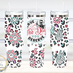 May include: A white stainless steel tumbler with a black handle. The tumbler has a white background with a leopard print pattern, pink and blue flowers, lightning bolts, and a skull and crossbones. The text "Have the day you deserve" is printed on the tumbler.