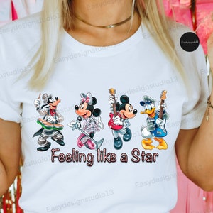 Pode incluir: Camiseta branca com um gráfico de Mickey Mouse, Minnie Mouse, Pateta e Donald Duck tocando instrumentos musicais. O texto "Feeling like a Star" está impresso abaixo do gráfico.
