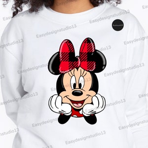 Pode incluir: Camisola branca com um desenho de desenho animado da Minnie Mouse. Minnie Mouse é retratada com orelhas pretas, um laço xadrez vermelho e preto e um vestido de bolinhas vermelho e branco. O desenho está centrado na frente da camisola.